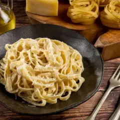 Talerz pełen tagliatelle z kremowym, oryginalnym sosem alfredo, obok butelka oliwy, ser i surowy makaron.