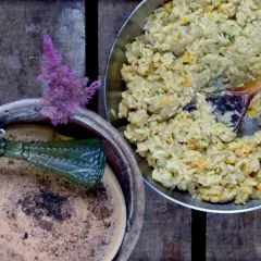 Kremowe risotto, jak z kuchni weneckiej, z drewnianą łyżką w metalowej patelni. Obok gliniana donica z zieloną butelką i fioletowym kwiatem.