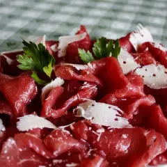 Cienkie plastry soczystej carne salada, posypane parmezanem i udekorowane listkami pietruszki, na zielono-białym obrusie.