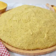 Duża, okrągła polenta bergamo na drewnianej desce, obok miseczka z mąką kukurydzianą i drewniana łopatka.