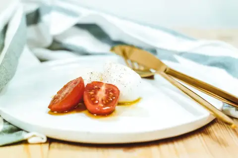Ser mozzarella di bufala z pomidorkami koktajlowymi na talerzu, obok złote sztućce i ściereczka.