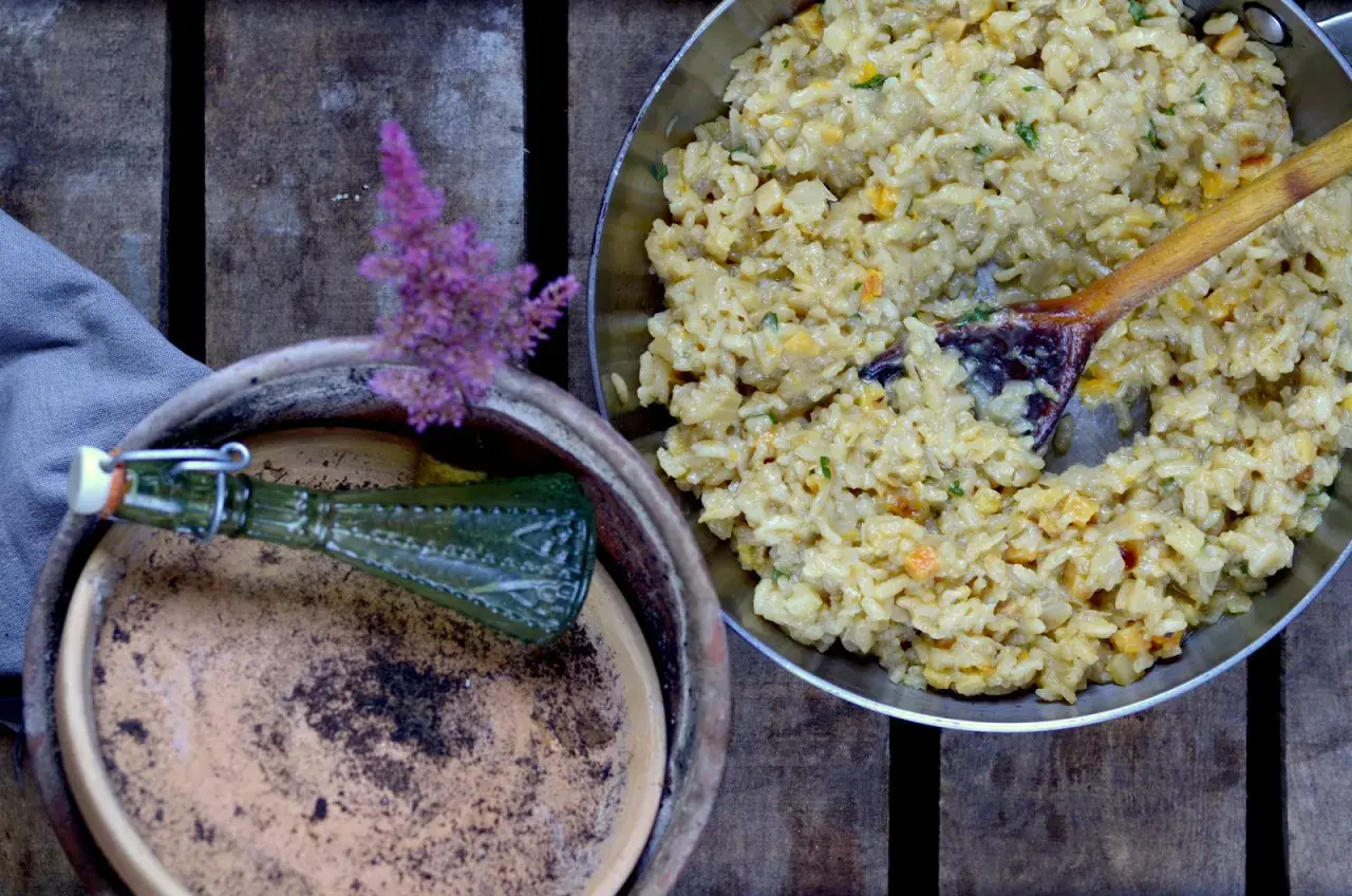 Kremowe risotto, jak z kuchni weneckiej, z drewnianą łyżką w metalowej patelni. Obok gliniana donica z zieloną butelką i fioletowym kwiatem.