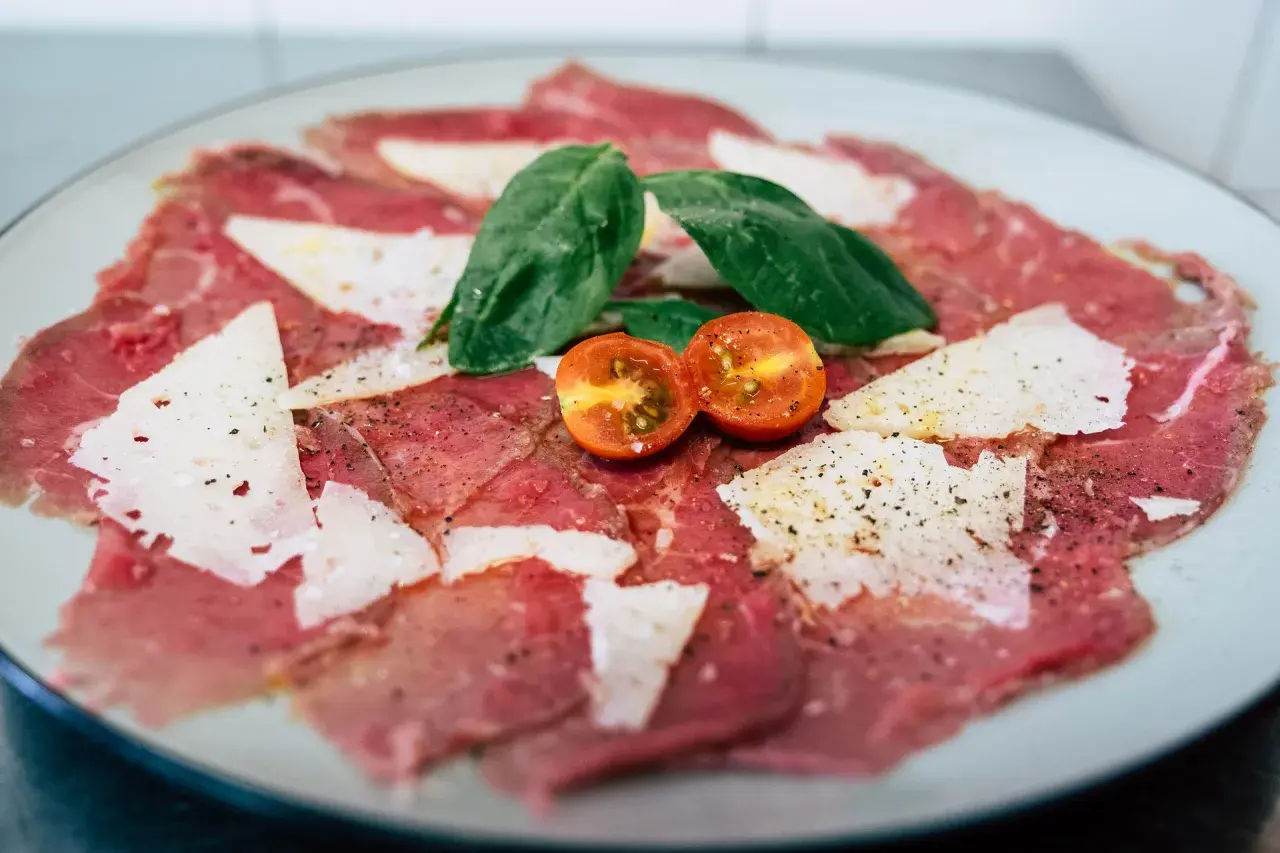 Eleganckie carpaccio wołowe z płatkami parmezanu, listkami bazylii i połówkami pomidorków koktajlowych, doprawione świeżo mielonym pieprzem.