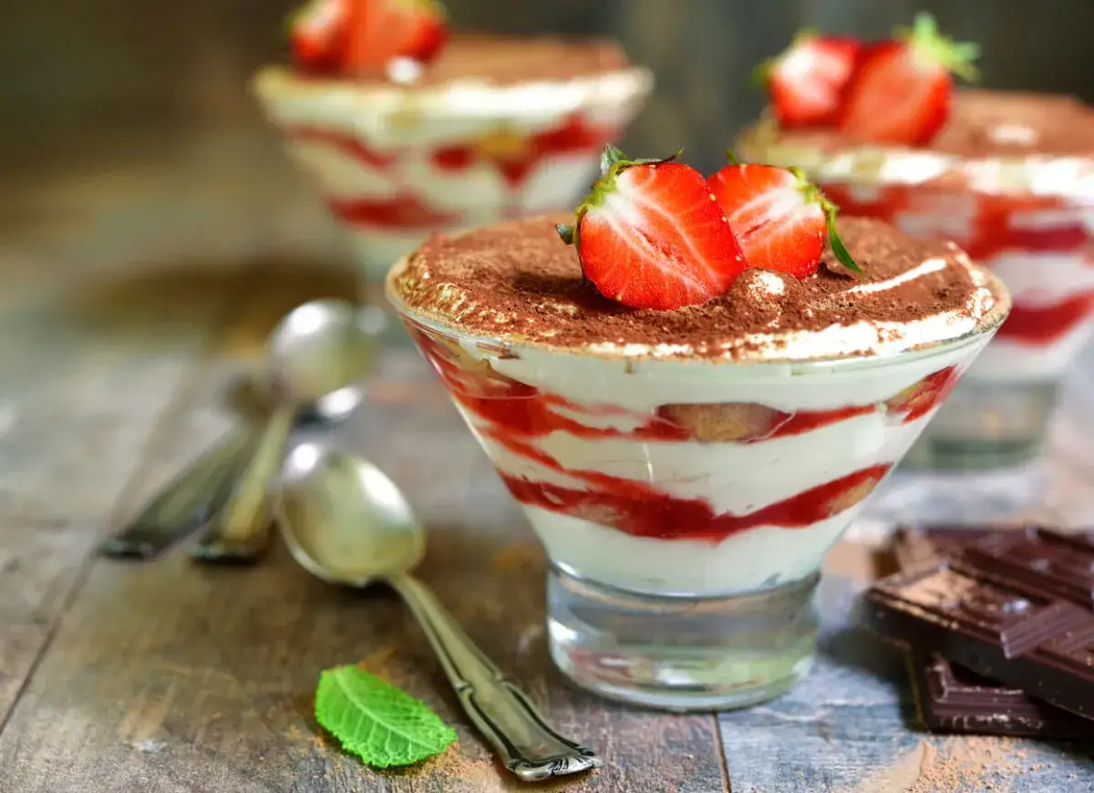Pyszne tiramisu z truskawkami i kremem z mascarpone, posypane kakao. Obok leżą łyżeczki i kawałki czekolady.